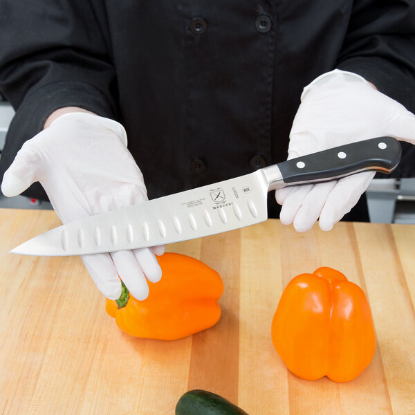 A hand in white gloves uses a Mercer Culinary Renaissance chef's knife to cut an orange bell pepper.