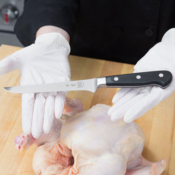 A gloved hand uses a Mercer Culinary Renaissance Stiff Boning Knife to cut raw chicken.