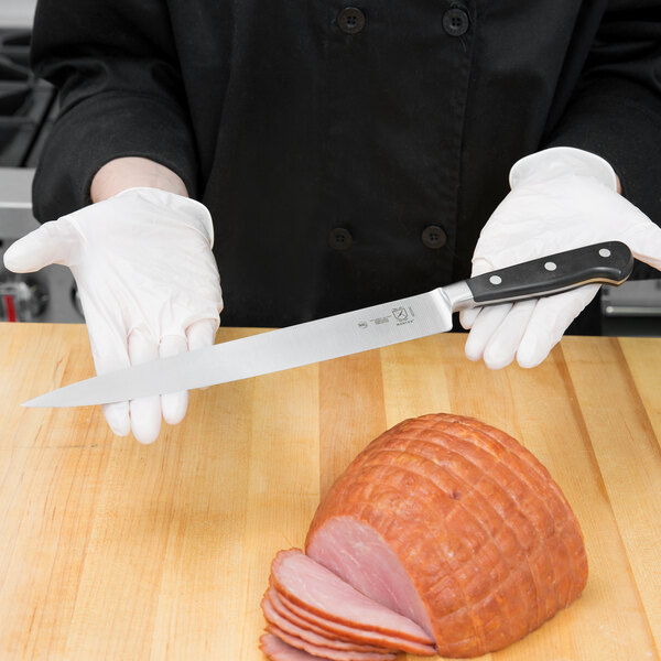 A person wearing white gloves uses a Mercer Culinary Renaissance Forged Carving Knife to cut a ham.
