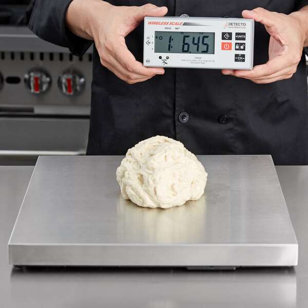 A person holding a Cardinal Detecto stainless steel pizza scale with a digital display over a ball of dough on a metal surface.