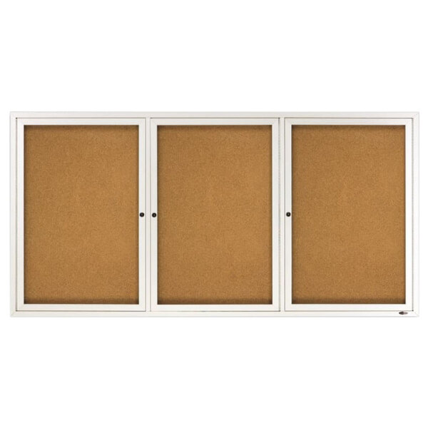 A silver aluminum enclosed bulletin board with a cork board inside and three white doors.