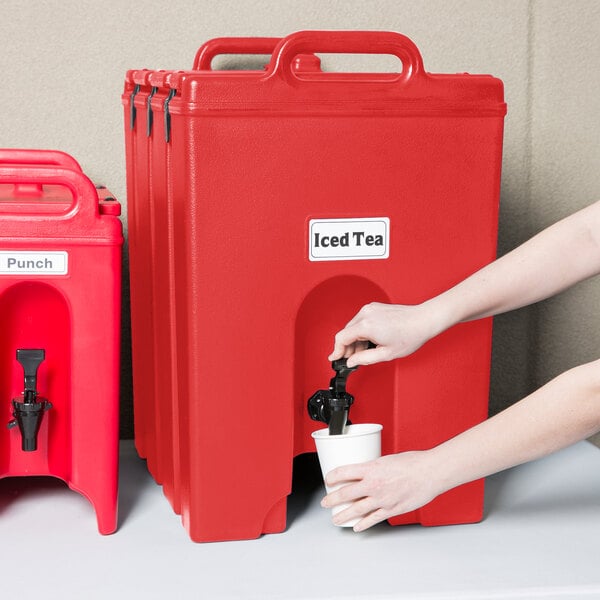 A person pouring water from a red Cambro beverage dispenser into a cup.