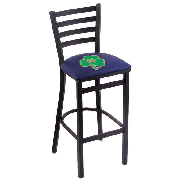 A blue bar height chair with a green and yellow clover logo on the padded seat and ladder back, with black steel legs.