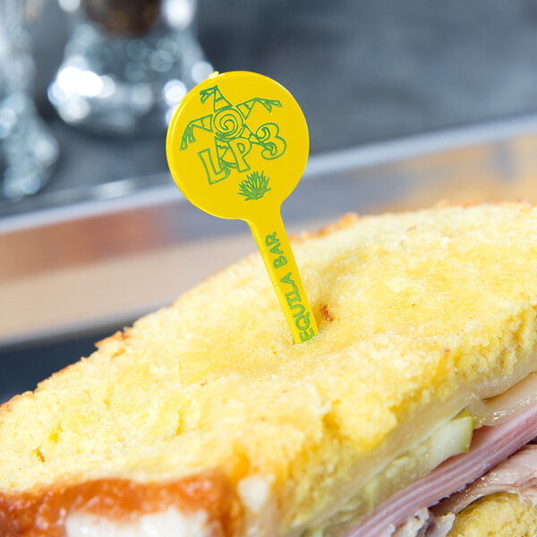 A sandwich with a yellow plastic toothpick featuring a yellow disc with writing on it.