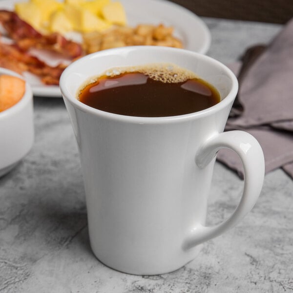 A white Libbey Flairique mug filled with brown liquid on a table with a plate of food and a cup of coffee.