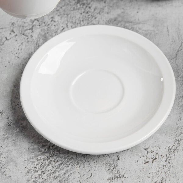 A Libbey bright white porcelain saucer holding a white cup on a marbled surface.