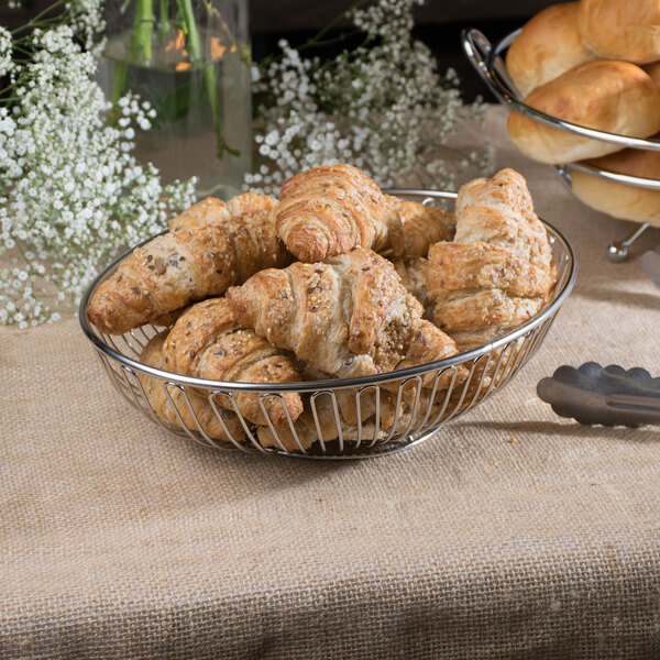 A Clipper Mill chrome plated iron oval wire basket filled with croissants on a table in a bakery display.