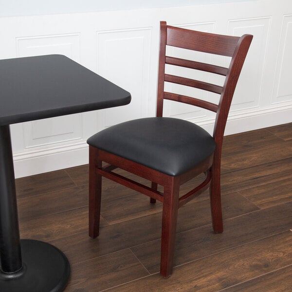 A Lancaster Table & Seating mahogany wood chair with a black vinyl seat next to a table in a restaurant.