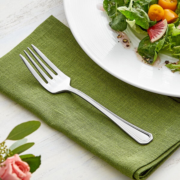 An Acopa stainless steel salad fork on a green napkin next to a plate with a salad.
