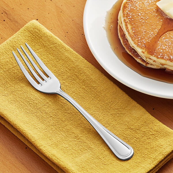 An Acopa Edgewood stainless steel dinner fork on a napkin next to a plate of pancakes.