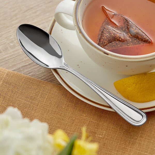 An Acopa stainless steel teaspoon on a saucer with a cup of tea and a tea bag.