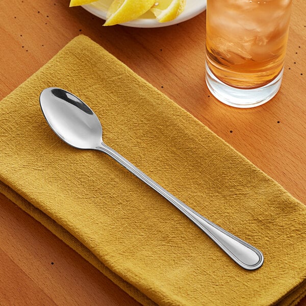 An Acopa Edgewood stainless steel iced tea spoon on a yellow napkin next to a glass of ice and a lemon.