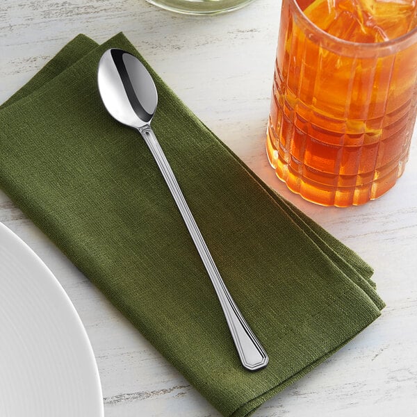 An Acopa stainless steel iced tea spoon on a green napkin next to a glass of orange liquid.
