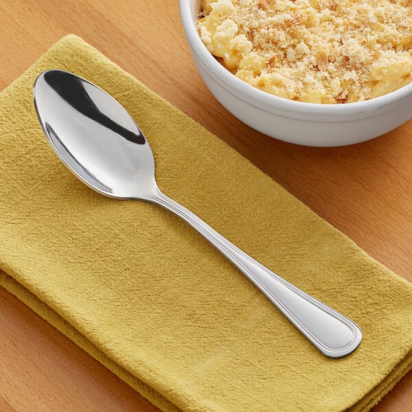 An Acopa Edgewood stainless steel serving spoon on a napkin next to a bowl of macaroni and cheese.