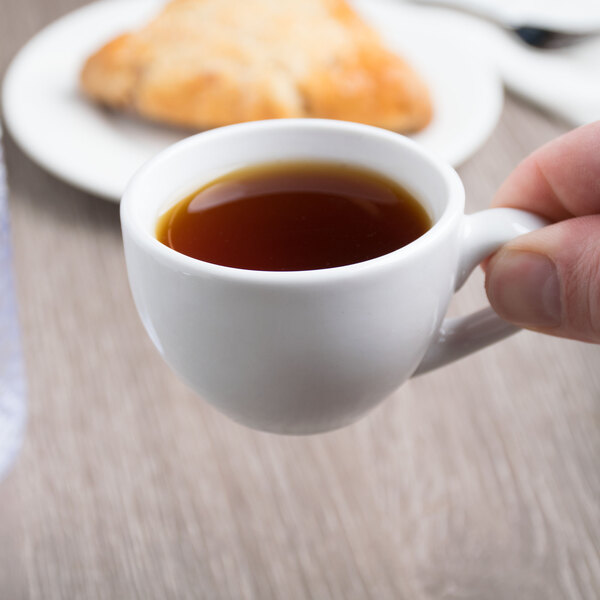 A person's hand holding a Libbey white porcelain espresso cup filled with brown coffee.