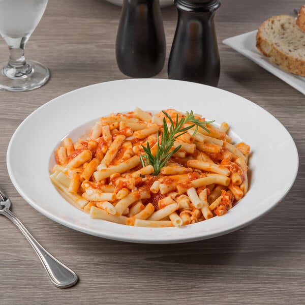 A Libbey porcelain bowl filled with pasta with sauce and rosemary on top on a table with a slice of bread.