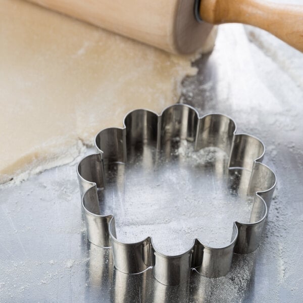 A metal Ateco dahlia flower cookie cutter on a table with a rolling pin.