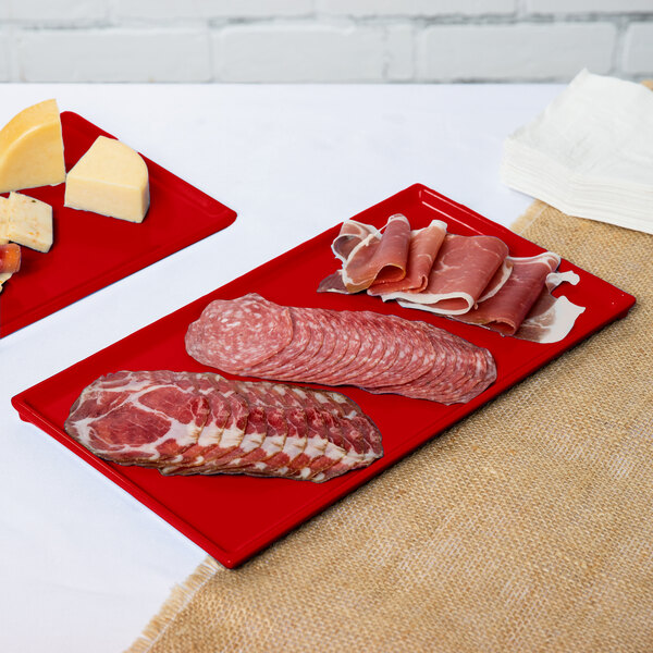 Two red Tablecraft cast aluminum rectangular cooling platters of meat and cheese on a counter.