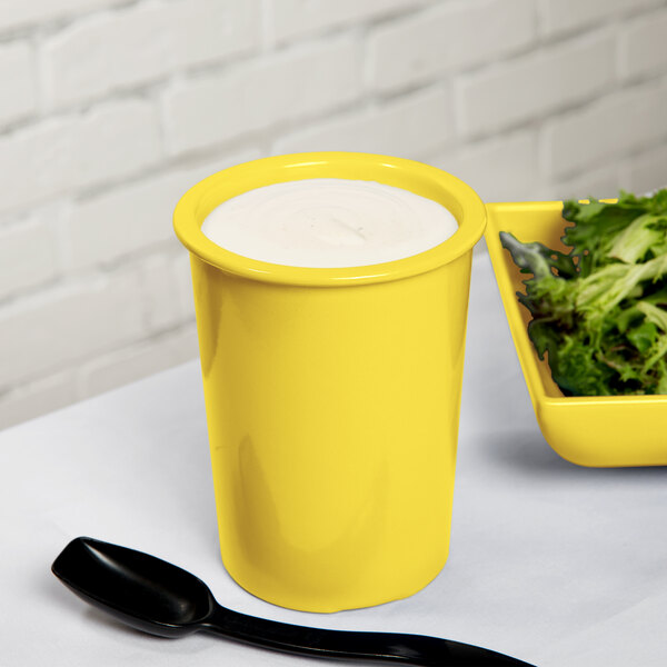 A close-up of a Tablecraft yellow cast aluminum salad dressing crock with white liquid in it next to a bowl of salad.