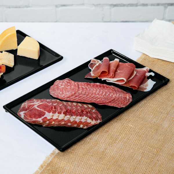 Two black Tablecraft rectangular metal trays with green speckles holding meat and cheese on a counter.