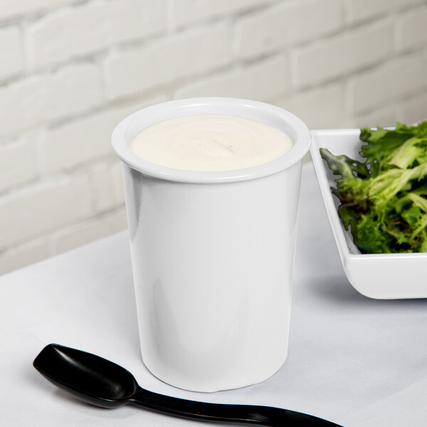 A Tablecraft white cast aluminum salad dressing crock on a table with a bowl of lettuce and a white cup of dressing.