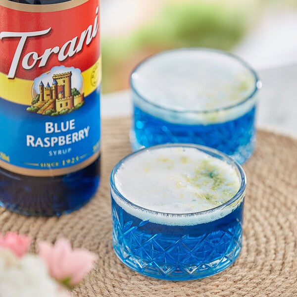 A blue drink in glasses next to a Torani Blue Raspberry Flavoring Syrup bottle.