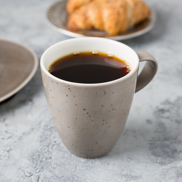 A Schonwald light gray porcelain cup filled with coffee next to a croissant.