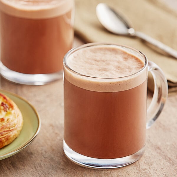 A glass mug of Ghirardelli sweet ground chocolate and cocoa with a pastry on a plate.