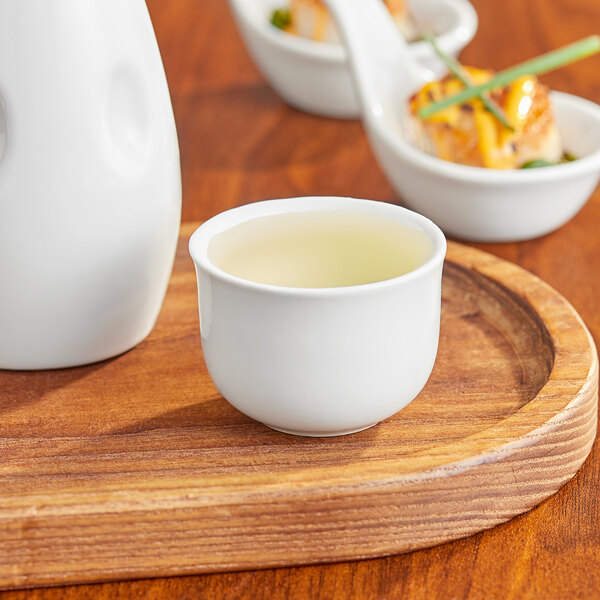 An Acopa bright white sake cup filled with liquid on a wooden tray.