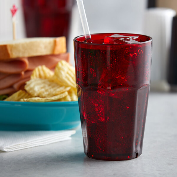 A red GET Bahama plastic tumbler with a straw and red liquid on a table with a sandwich and bowl of chips.