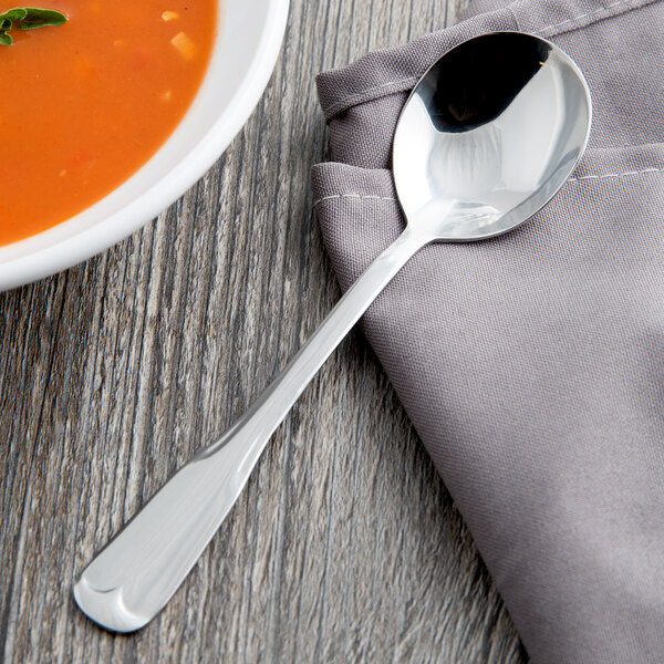 A World Tableware stainless steel bouillon spoon on a napkin next to a bowl of soup.