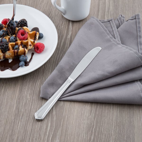 A Libbey Cortland stainless steel dessert knife on a napkin next to a plate of waffles with berries and chocolate sauce.