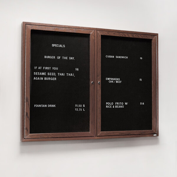 An Aarco walnut frame bulletin board with black felt and hinged doors on a wall with a black and white menu.