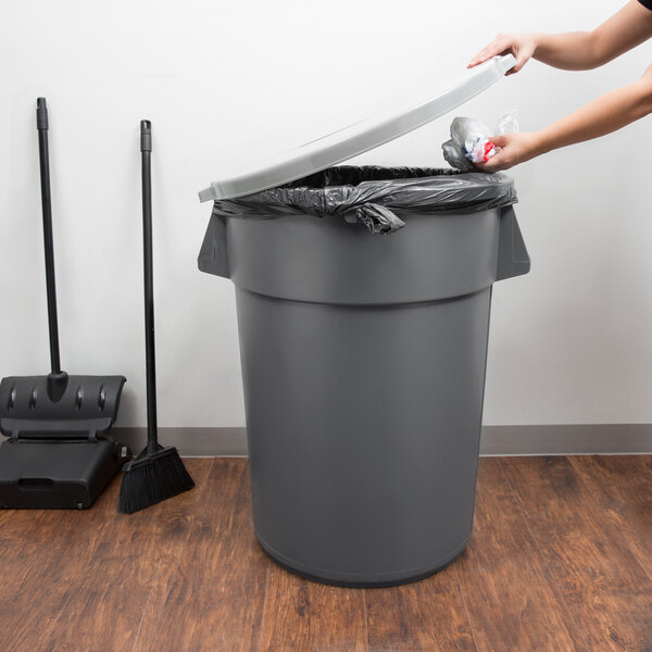 A woman using a broom to put a plastic bag in a grey Continental round trash can.