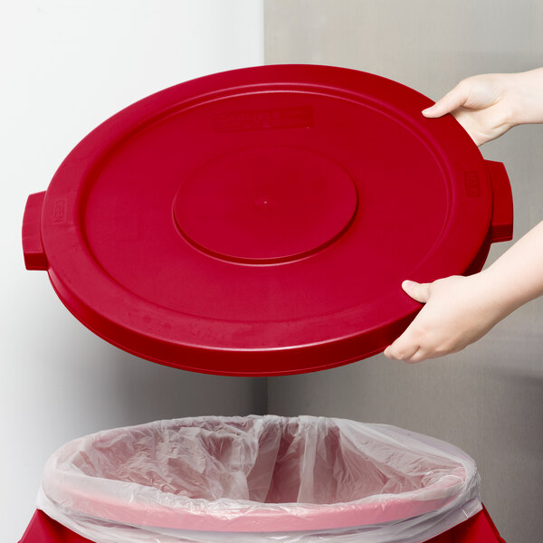 A person holding a red lid over a red trash can.