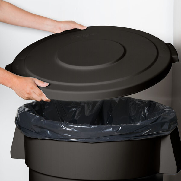 A hand holding a Carlisle black round trash can lid over a black garbage can.