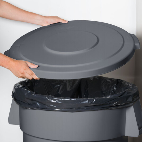 A person's hand holding a grey circular Carlisle Bronco trash can lid over a trash can.
