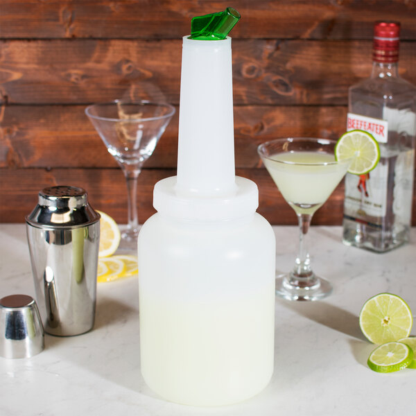 A white Carlisle Store 'N Pour container with a green lid next to a glass of liquid with a lime wedge in it.