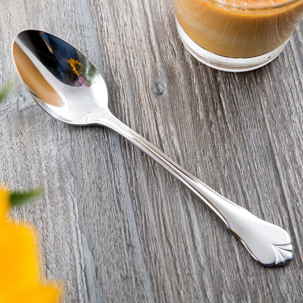 A Libbey stainless steel teaspoon with a flower design on the handle next to a glass of liquid on a table.