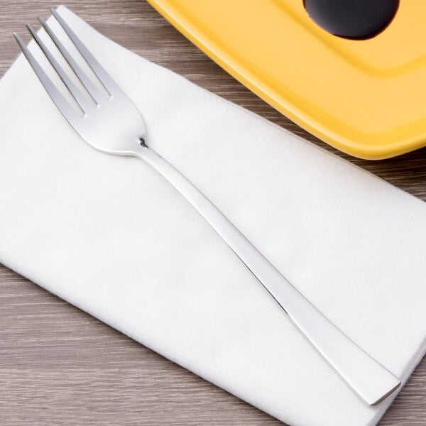 An Arcoroc stainless steel dinner fork on a white napkin next to a yellow plate.