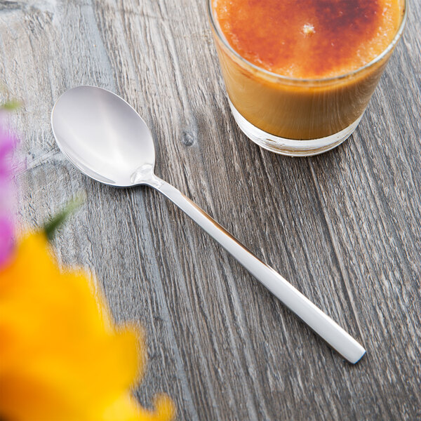 A Libbey Elexa stainless steel teaspoon on a table next to a glass of liquid.