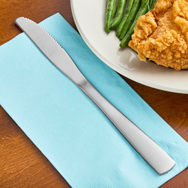 A Choice stainless steel medium weight dinner knife on a napkin next to a plate of fried chicken and green beans.