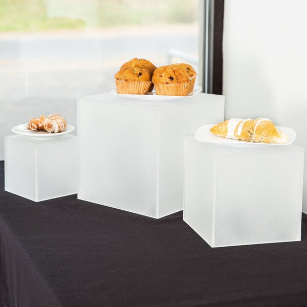 Three American Metalcraft frosted acrylic cube risers with plates of muffins on them.