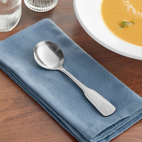 An Acopa stainless steel bouillon spoon on a blue napkin next to a bowl of soup.