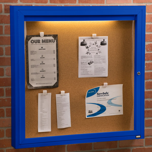 An Aarco blue indoor lighted bulletin board cabinet with a cork board inside with paper pinned to it.