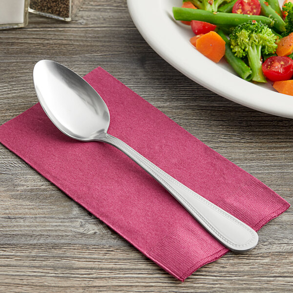 A Choice Milton stainless steel serving spoon on a napkin next to a plate of vegetables.