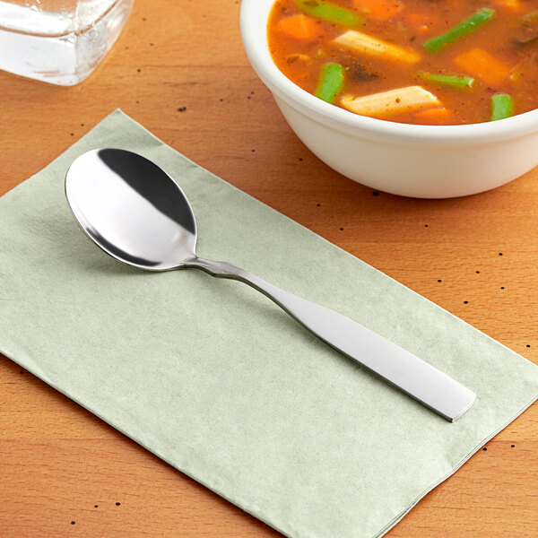 A Choice medium weight stainless steel bouillon spoon on a napkin next to a bowl of soup.