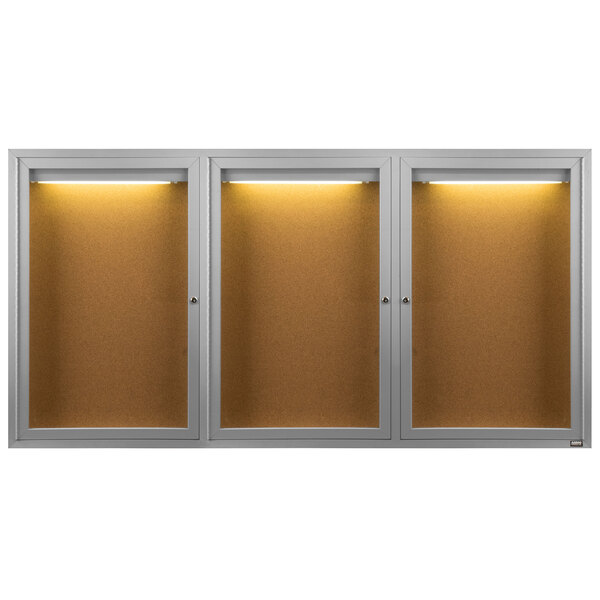 An Aarco satin anodized indoor bulletin board cabinet with three glass doors and lights.