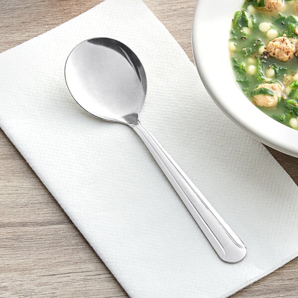 A Choice Dominion stainless steel bouillon spoon on a napkin next to a bowl of soup.