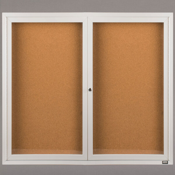 A white framed Aarco bulletin board cabinet with two glass doors showcasing a cork board inside.